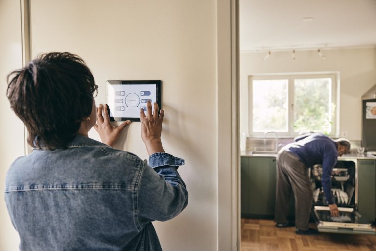 Senior woman wearing denim jacket using home automation system on wall while man in background loads dishwasher