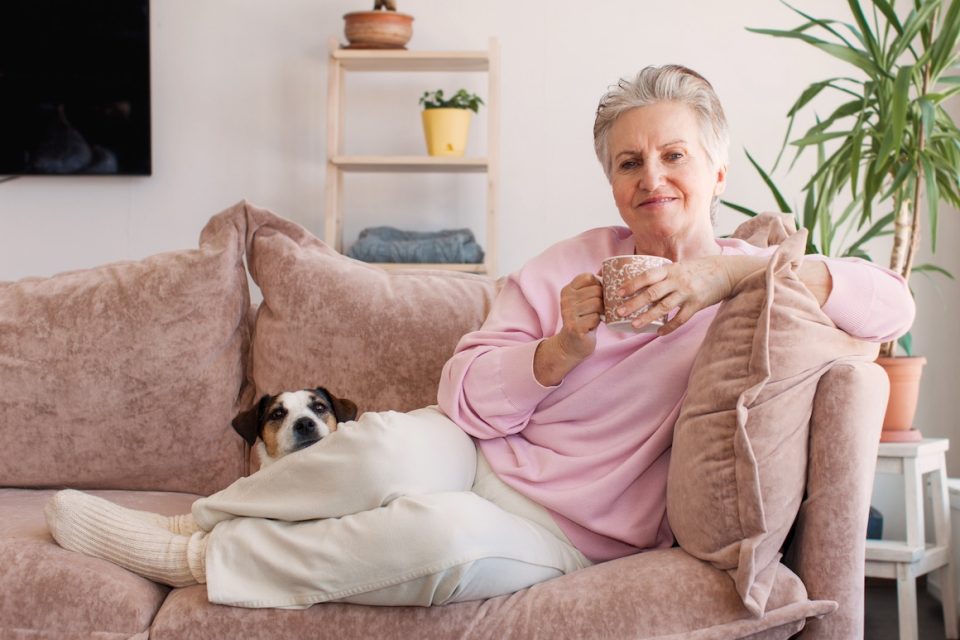 mature older adult grey-haired woman drinking coffee relaxing on sofa at home with dog